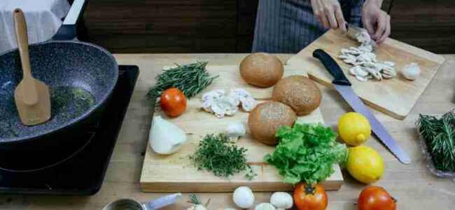 Crop anonymous female chef wearing apron cutting fresh mushrooms while standing at table with scattered ripe vegetables including onions tomatoes and potatoes in modern kitchen, tags: für öl - pexel