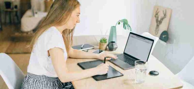 A young woman working remotely at a wooden table with her laptop and tablet in a cozy home setting., tags: arbeit und deutschland - pexel